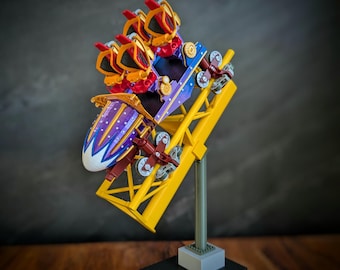 May include: A miniature roller coaster model with a yellow track, a purple and white car, and red and gold accents. The coaster is mounted on a gray and black base, set against a dark background. The model is a detailed representation of a theme park ride.