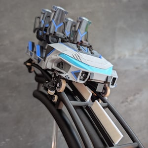 Könnte beinhalten: Ein Miniatur-Achterbahnwagen-Modell mit weißer und hellblauer Außenhülle und schwarzen Akzenten. Der Wagen verfügt über vier Sitze mit schwarzen Kopfstützen und blauen Details und ist auf einem schwarzen Schienenabschnitt mit kleinen Rädern montiert.