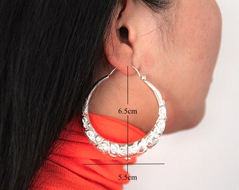May include: Silver hoop earrings with an ornate floral design. The earrings measure 6.5 cm in diameter and 5.5 cm in width. The earrings are shown on a person's ear, with black hair and an orange top in the background.