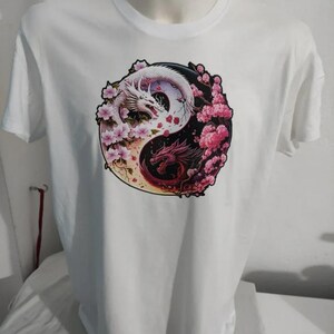 Puede incluir: Camiseta blanca con un diseño de Yin Yang con dos dragones. Un dragón es blanco, el otro rojo, rodeados de flores de cerezo rosas. La camiseta se muestra en un maniquí.
