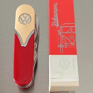 Op de afbeelding: Een rood en goudkleurig multifunctioneel zakmes met een zilveren Volkswagen-logo op het handvat. Daarnaast staat een rechthoekige rood-witte doos met de tekst Volkswagen, een lijntekening van een busje en een Official Product-sticker met logo.