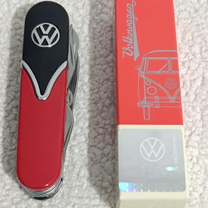 Può includere: Un coltellino multiuso con manico rosso e nero caratterizzato dal logo Volkswagen argentato, posizionato accanto a una scatola rettangolare rossa e bianca. La scatola mostra la scritta Volkswagen, un disegno al tratto di un furgone e un adesivo olografico.