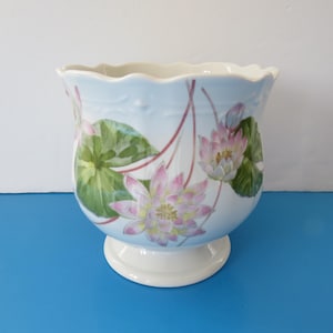 Peut inclure: Un cache-pot en porcelaine blanche avec un bord festonné et une base sur pied. L'extérieur présente un fond bleu clair orné de fleurs de lotus roses peintes, de nénuphars verts et de fines tiges brun-rouge entourant le corps.