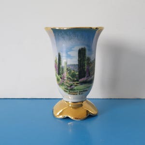 May include: A small decorative vase with a flared top and a gold base. The vase has a landscape painting with trees, flowers, and water. The rim and base are gold. Text on the vase reads "The Butchart Gardens, Victoria, B.C."