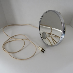 May include: A round, chrome-framed electric vanity mirror with a reflective surface and a small circular magnifying inset. The mirror features a long, beige power cord with a standard two-prong electrical plug attached to the side of the frame.