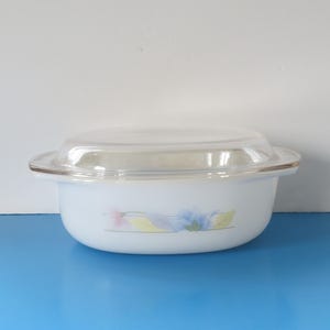 Peut inclure: Plat à casserole rond blanc avec un couvercle en verre transparent. Le plat présente un motif floral aux couleurs pastel. Le plat est posé sur une surface bleue.