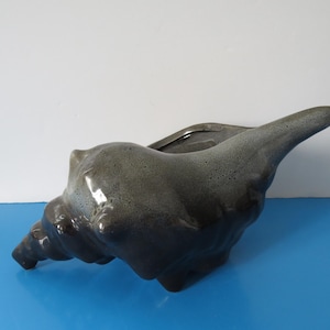 Peut inclure: Un pot de fleurs en céramique en forme de coquillage, dans des tons de gris et de noir. Le pot a une finition brillante et une surface texturée, avec une large ouverture pour les plantes ou les objets décoratifs. Le design imite une coquille naturelle.