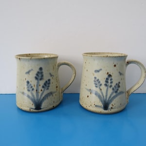 May include: Two ceramic mugs with a speckled beige finish. Each mug features a blue floral design and small bird accents. The mugs have a curved handle and are set against a blue surface and white background.