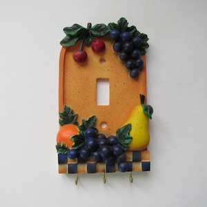 Peut inclure: Plaque décorative pour interrupteur avec un thème de fruits. La plaque est de couleur orange clair avec une surface texturée. Elle présente une ouverture pour l'interrupteur, une grappe de raisins, une poire, des cerises et une orange. Le bas a un motif à carreaux et trois petits crochets.
