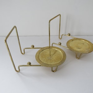 May include: Two vintage brass candle holders. Each holder features a round, etched plate supported by three small legs. The plates are connected to a vertical, angular frame. The brass has a warm, aged patina, suggesting a mid-century aesthetic.