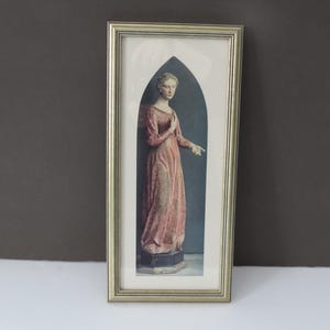 Peut inclure: Une estampe encadrée d'une peinture représentant une femme en robe longue rose debout dans une arche pointue. La femme regarde vers la droite et sa main droite est levée.