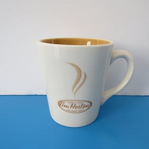 Peut inclure: Mug à café en céramique blanche avec un intérieur marron et une anse incurvée. La tasse présente un motif de vapeur stylisé et le logo Tim Hortons. La tasse est posée sur une surface bleue.