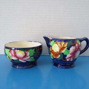 Peut inclure: Un ensemble de crémier et sucrier en céramique bleue. Les pièces sont décorées de grandes fleurs roses, jaunes et marron sur fond bleu foncé. Le sucrier est rond et le crémier a une anse.