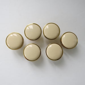 Peut inclure: Six boutons de meuble crème avec une bordure dorée. Les boutons sont ronds et ont une finition brillante. Ces boutons décoratifs conviennent à la restauration de meubles ou aux projets de bricolage.
