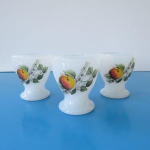 Peut inclure: Trois coquetiers blancs ornés d'un motif de pomme rouge et jaune, de feuilles vertes et de branches de bouleau. Les coquetiers sont posés sur une surface bleue.