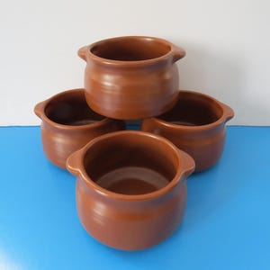 Peut inclure: Quatre bols en céramique de couleur terre cuite empilés les uns sur les autres. Les bols ont une forme arrondie et un bord légèrement évasé.