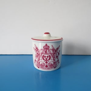 May include: A white ceramic jar with a lid, featuring a red crest design and the words "QUE MAILLE QUIMAILLE". The jar has a red rim and a red knob on the lid. The jar is on a blue surface.