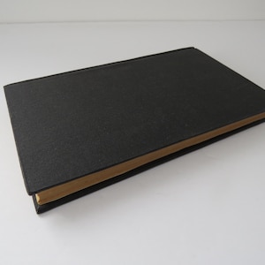 Peut inclure: Un livre fermé à couverture rigide noire et pages beiges. Le livre est posé à plat sur une surface blanche. La couverture a un aspect texturé.