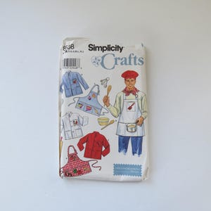 Peut inclure: Une pochette de patron de couture Simplicity Crafts, numéro 8508, présentant des illustrations de tabliers et de vestes. La couverture montre un chef portant un tablier blanc et une toque rouge, ainsi que divers modèles de vêtements.
