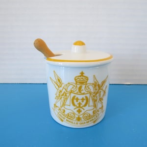 May include: A white ceramic jar with a lid and a wooden spoon. The jar has a yellow rim and a yellow emblem with the words "QUE MAILLE QUI MAILLE". The jar is on a blue surface.
