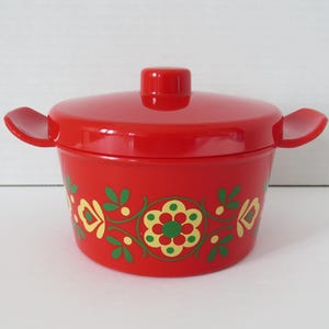 Peut inclure: Un plat à casserole rouge avec un couvercle et des poignées latérales. Le plat est orné d'un motif floral vert, jaune et rouge. Le couvercle a un petit bouton. Le plat est probablement en plastique ou en céramique.