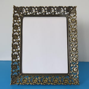 Peut inclure: Un cadre photo rectangulaire orné avec une finition bronze. Le cadre est décoré d'un motif de roses répété sur les bords. Le cadre est vide, avec un fond blanc visible à l'intérieur.