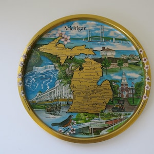May include: A round metal decorative tray with a gold rim featuring a map of Michigan. The illustration includes the state name, landmarks like the Grand Hotel, a bridge, a ship, a bird, and floral accents along the border on a blue background.