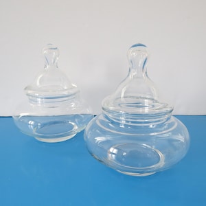 May include: Two clear glass jars with lids. The jars are round and have a wide mouth. The lids are domed and have a knob on top.
