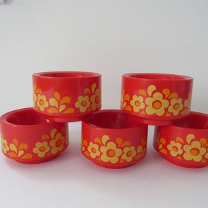 May include: Five red, cylindrical containers with a floral pattern in yellow and orange. The containers are stacked, showcasing their design. The floral pattern is repeated around the circumference of each container.