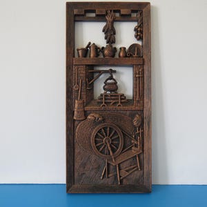 May include: A brown wooden wall hanging featuring a carved relief of a rustic kitchen scene. The artwork includes a spinning wheel, a pot over a fire, and various kitchen utensils. The frame is rectangular with a detailed border.