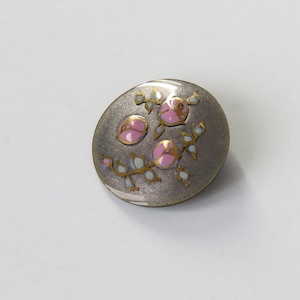 May include: An oval silver button with a floral pattern. The button has three pink rose motifs with gold detailing and pale blue leaves. The button is set against a white backdrop.