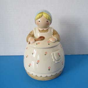 May include: A ceramic cookie jar in the shape of a woman wearing a blue bonnet and apron. The off-white apron is decorated with small pink flowers. The woman is holding a wooden spoon.