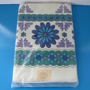 Peut inclure: Une nappe florale vintage pliée dans un emballage plastique. La nappe présente une grande fleur bleue au centre, entourée de petites fleurs et de motifs en forme de feuilles bleues, vertes et violettes sur fond blanc.