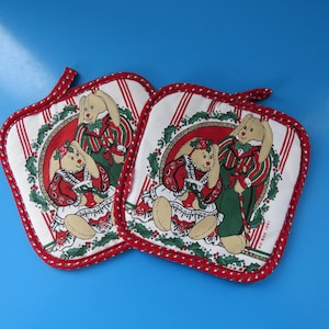 Peut inclure: Deux maniques carrées avec un motif de lapin de Noël. Chaque manique a un fond blanc avec des rayures rouges et blanches et une bordure rouge. Le centre présente un couple de lapins en tenue de fête.