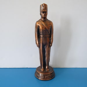 Peut inclure: Statue de soldat en bronze, portant un uniforme formel, comprenant un chapeau, une veste et un pantalon. La statue est sur une base ronde avec l'inscription "WEST POINT". La statue mesure environ 30 cm de haut.