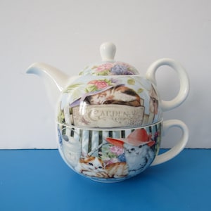 May include: A white ceramic teapot and teacup set with a whimsical cat garden design. The teapot features a sleeping cat, while the teacup showcases kittens wearing hats. Floral and striped patterns adorn the set, creating a charming aesthetic.