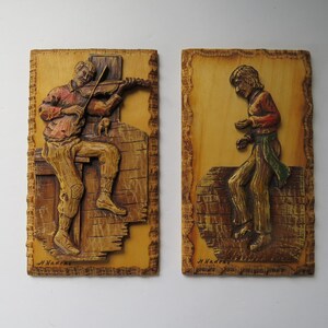 Peut inclure: Deux sculptures en relief en bois. L'une représente une figure assise jouant du violon, l'autre une figure debout. Les deux sont sculptées dans un style rustique avec des tons bruns et beiges chauds. Les sculptures sont montées sur des plaques de bois rectangulaires.