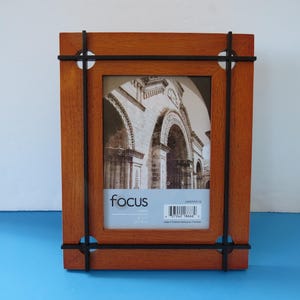 May include: A rectangular wooden picture frame with a medium brown finish, accented by a black metal frame overlay that extends past the edges. The frame contains a display insert featuring a black and white image of an archway and the text focus.