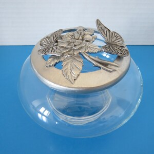 Peut inclure: Un bocal en verre transparent avec un couvercle décoratif argenté. Le couvercle présente un motif floral avec deux papillons. Le bocal est rond et repose sur une surface bleue. Le couvercle comprend une rose et des feuilles.
