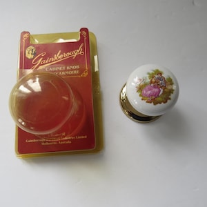 Peut inclure: Un bouton de porte de placard en céramique blanche avec une base dorée et un motif floral représentant un couple dans un jardin. Le bouton est emballé avec une étiquette Gainsborough Cabinet Knob & Armour.