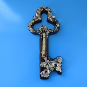 May include: Black key-shaped thermometer with the text "SAN DIEGO". The key is decorated with small, colorful shells and has a thermometer in the center. The background is a solid blue.