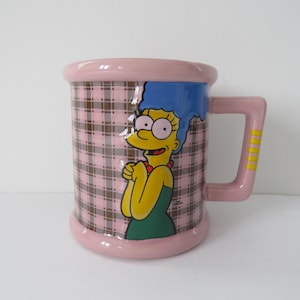May include: A ceramic mug featuring Marge Simpson from The Simpsons. The mug has a pink rim, handle, and base, with a plaid pattern on the body. Marge is depicted in her signature yellow skin, blue hair, and green dress.
