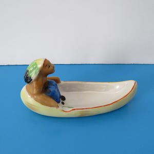May include: A ceramic figurine of a Native American person in a canoe. The canoe is yellow and white with red and green accents. The figurine is painted in shades of brown, blue, green, and yellow.