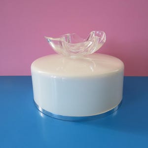 May include: A white, cylindrical decorative object with a silver base, topped with a clear, organically shaped glass bowl. The background is a vibrant combination of pink and blue.