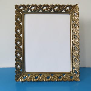 May include: An antique-style picture frame in a gold colour, with a rectangular aperture. The frame has an elaborate, scrolling leaf pattern around the border. It sits on a blue surface, with a white backdrop.