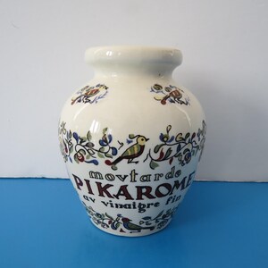 Peut inclure: Pot en céramique crème avec un corps arrondi et un col étroit. Le pot est décoré de motifs colorés d'oiseaux et de fleurs. Le texte sur le pot indique "movtarde PIKAROME av vinaigre fin".