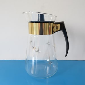 May include: A clear glass pitcher featuring a gold-toned metal band around the top, a black plastic handle, and a black lid. The glass body is decorated with a repeating pattern of gold-colored starburst designs scattered across the exterior surface.