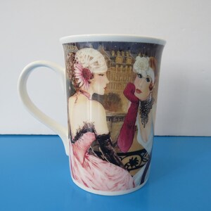 May include: White ceramic mug featuring an illustration of two women in vintage attire. One woman wears a pink dress and the other a white dress with red gloves. The background depicts a cityscape.