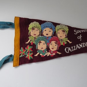 May include: A burgundy felt pennant with a gold border and teal ties. It features five figures in bonnets and the text "Souvenir of Callander". A decorative souvenir.