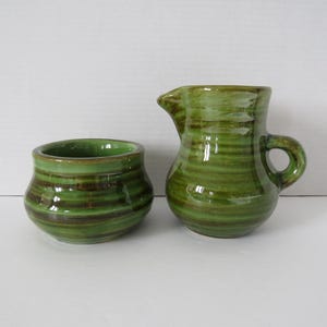 Peut inclure: Un pot à lait en céramique et un sucrier assorti présentant une glaçure brillante aux tons verts et bruns nuancés avec des textures striées horizontales. Le pot possède une anse courbée et un bec verseur, tandis que le bol a une ouverture large.